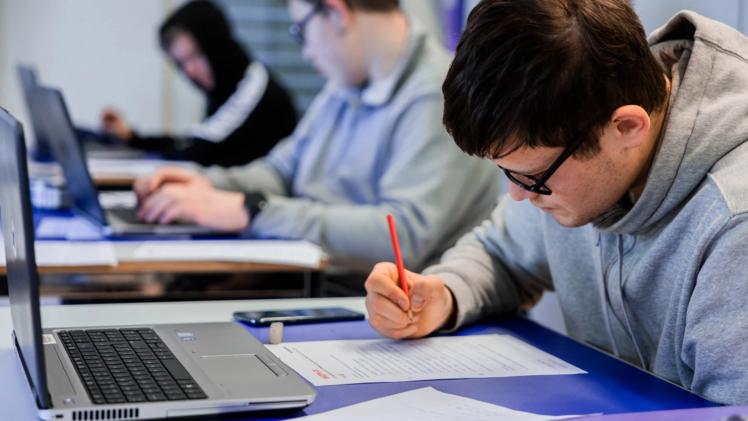Drei junge Auszubildende lernen an ihren Computern, einer von ihnen schreibt auf ein Blatt.